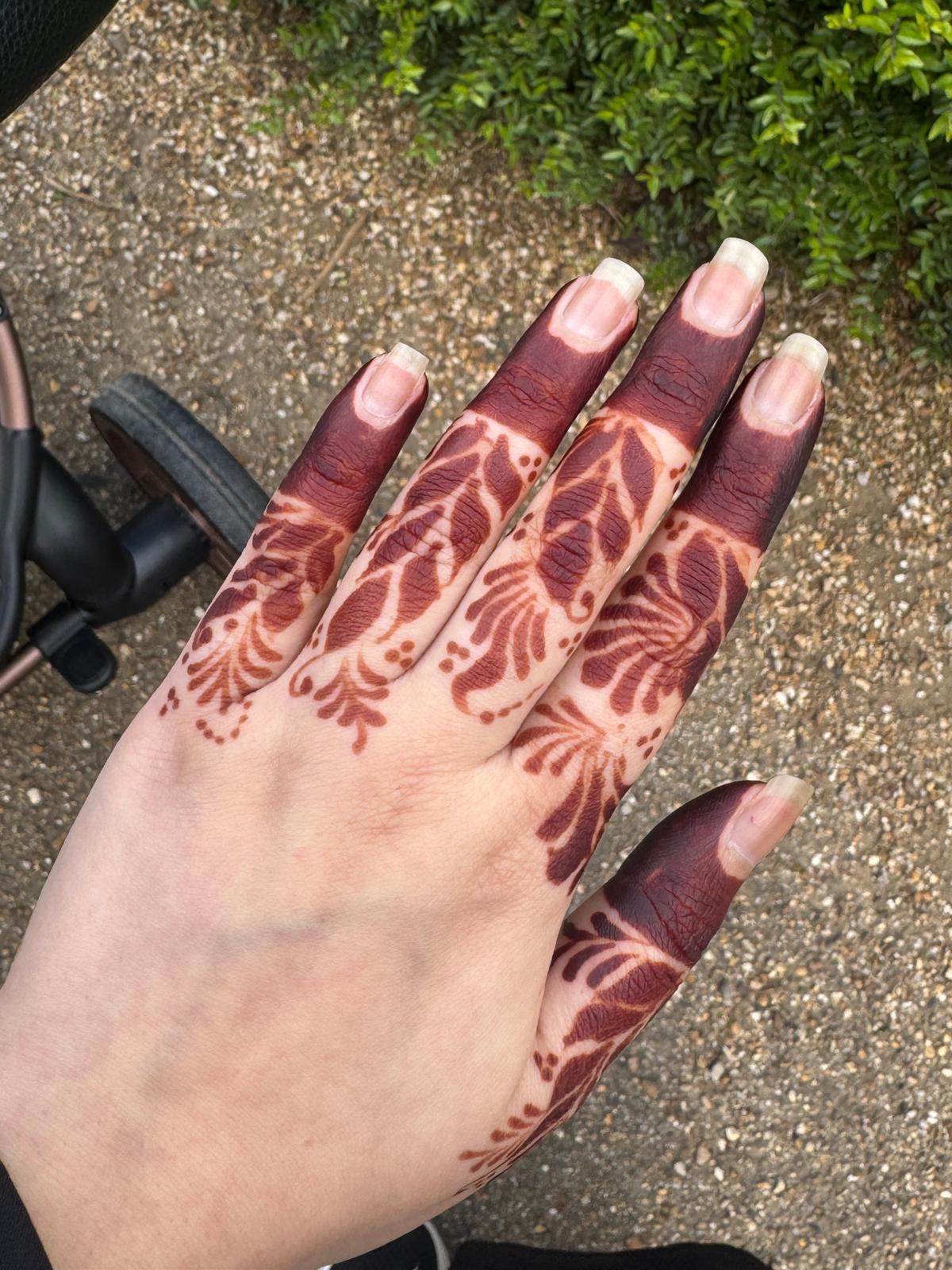 A close-up of a hand with intricate brown henna or mehndi patterns applied across the palm and fingers, resting on a dark gray textured fabric. A ring is worn on one finger.
