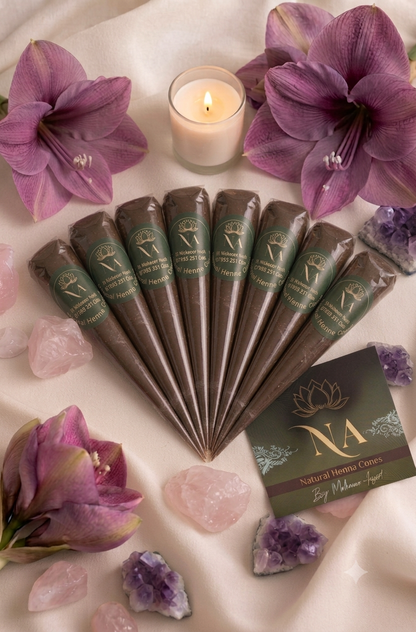 A set of brown cone-shaped incense sticks with green labels arranged in a fan shape on a draped cream fabric, surrounded by purple amaryllis, pink rose quartz, purple amethyst crystals, and a lit candle. A branded card for 'NA Natural Home Cares' is visible in the lower right.