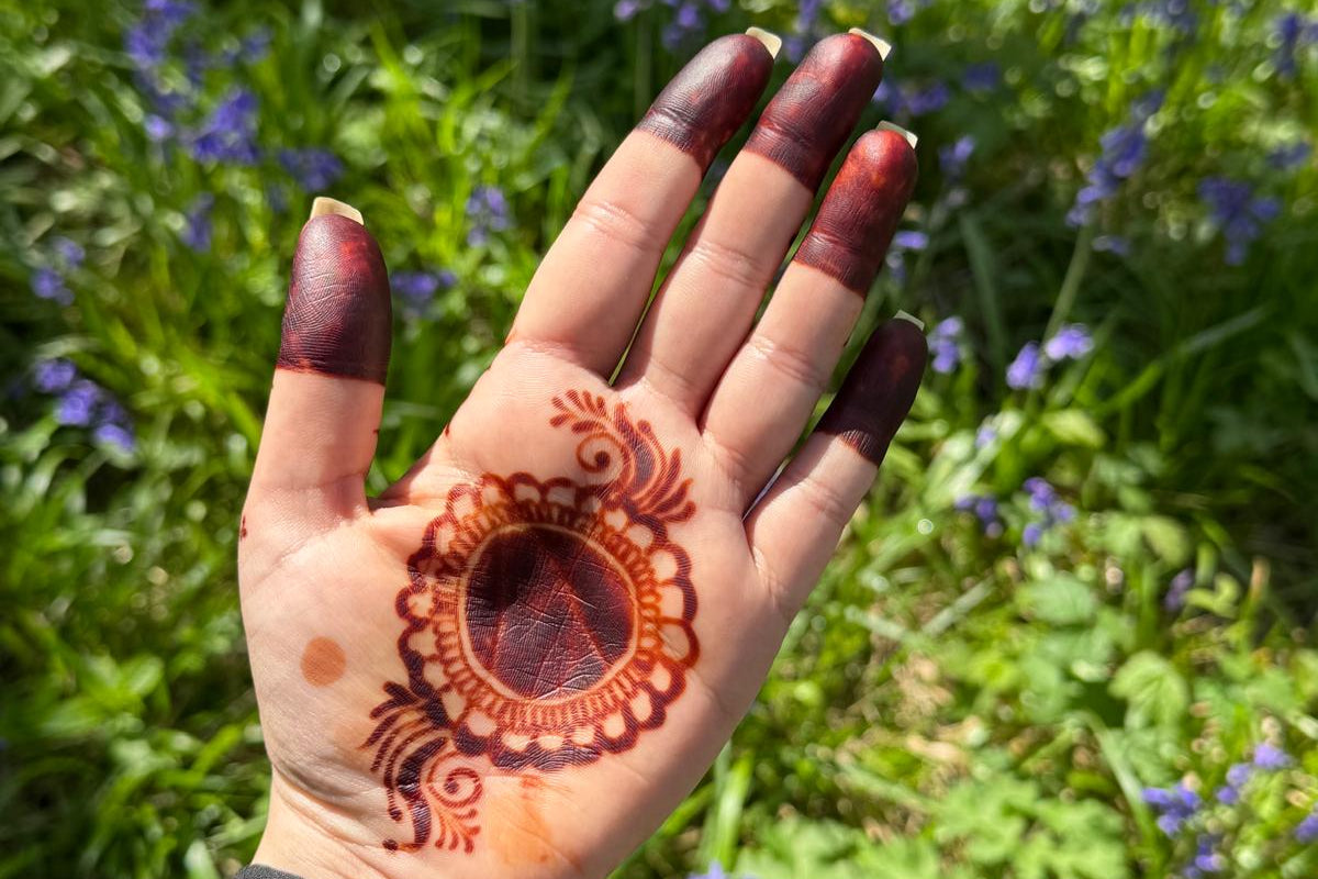A close-up of a hand with a detailed henna design on the palm and dark red painted fingernails, set against a blurred background of green grass and small purple flowers.