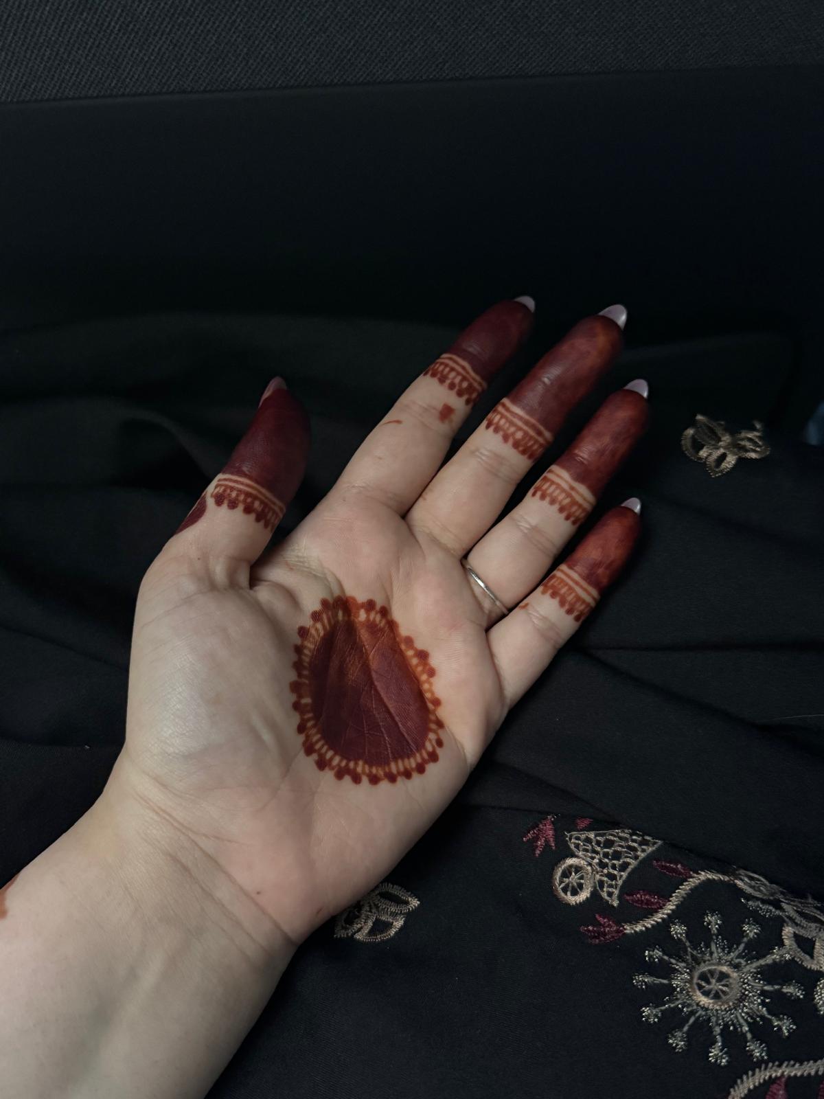 A close-up of a hand with intricate dark red henna designs on the palm and fingers, resting on a black embroidered fabric surface.