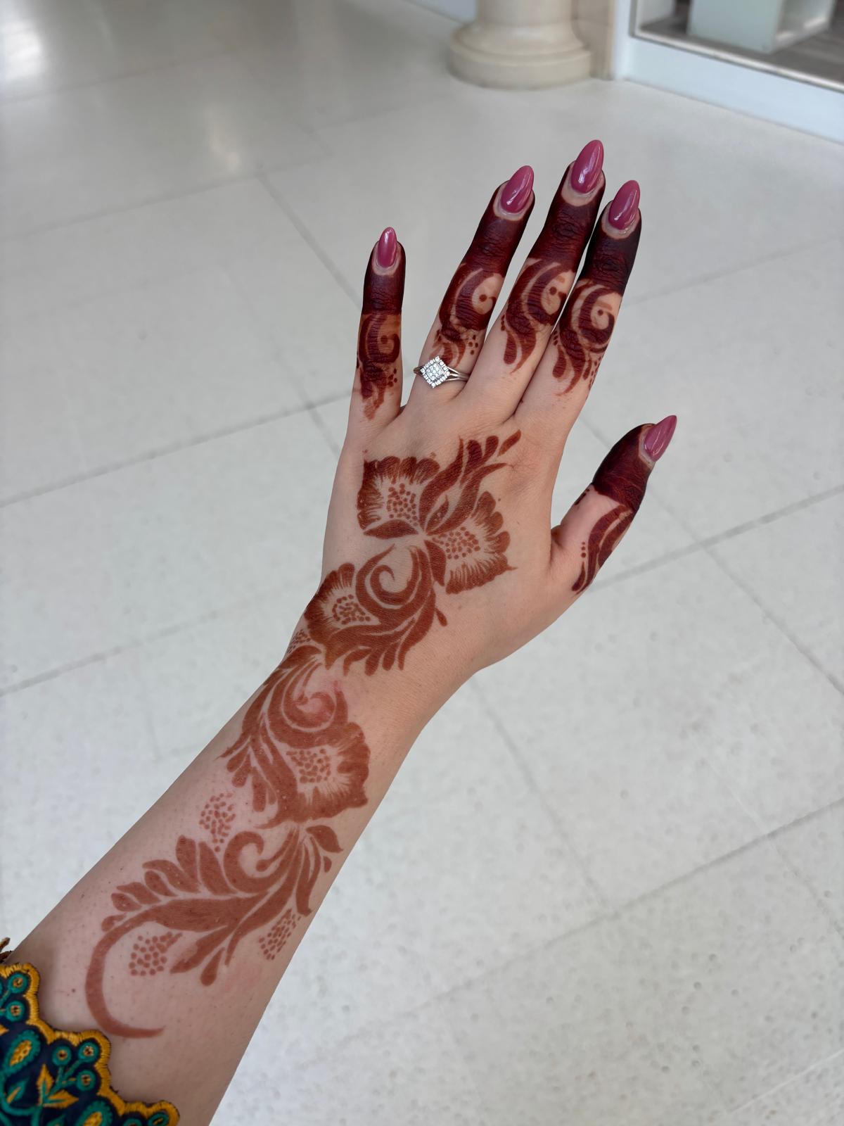 A hand and wrist decorated with intricate dark brown henna designs, featuring long nails with pink polish and a silver ring, set against a light tiled floor background.