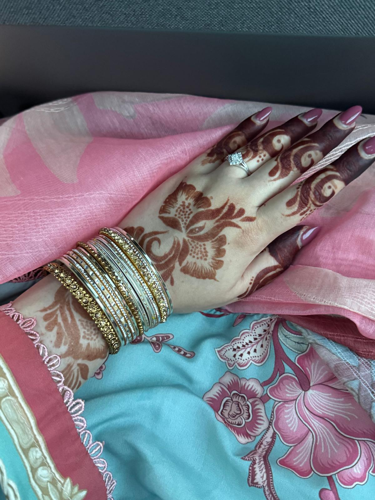 A close-up of a hand with intricate brown henna or mehndi patterns applied across the palm and fingers, resting on a dark gray textured fabric. A ring is worn on one finger.

