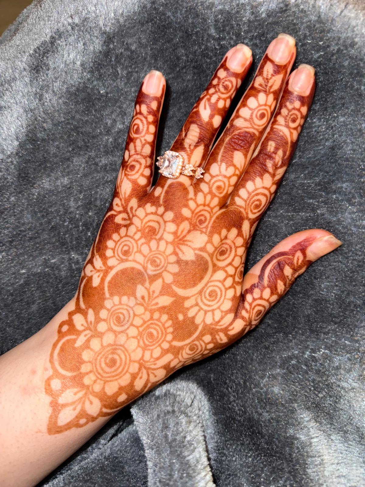 A close-up of a hand with intricate brown henna or mehndi patterns applied across the palm and fingers, resting on a dark gray textured fabric. A ring is worn on one finger.
