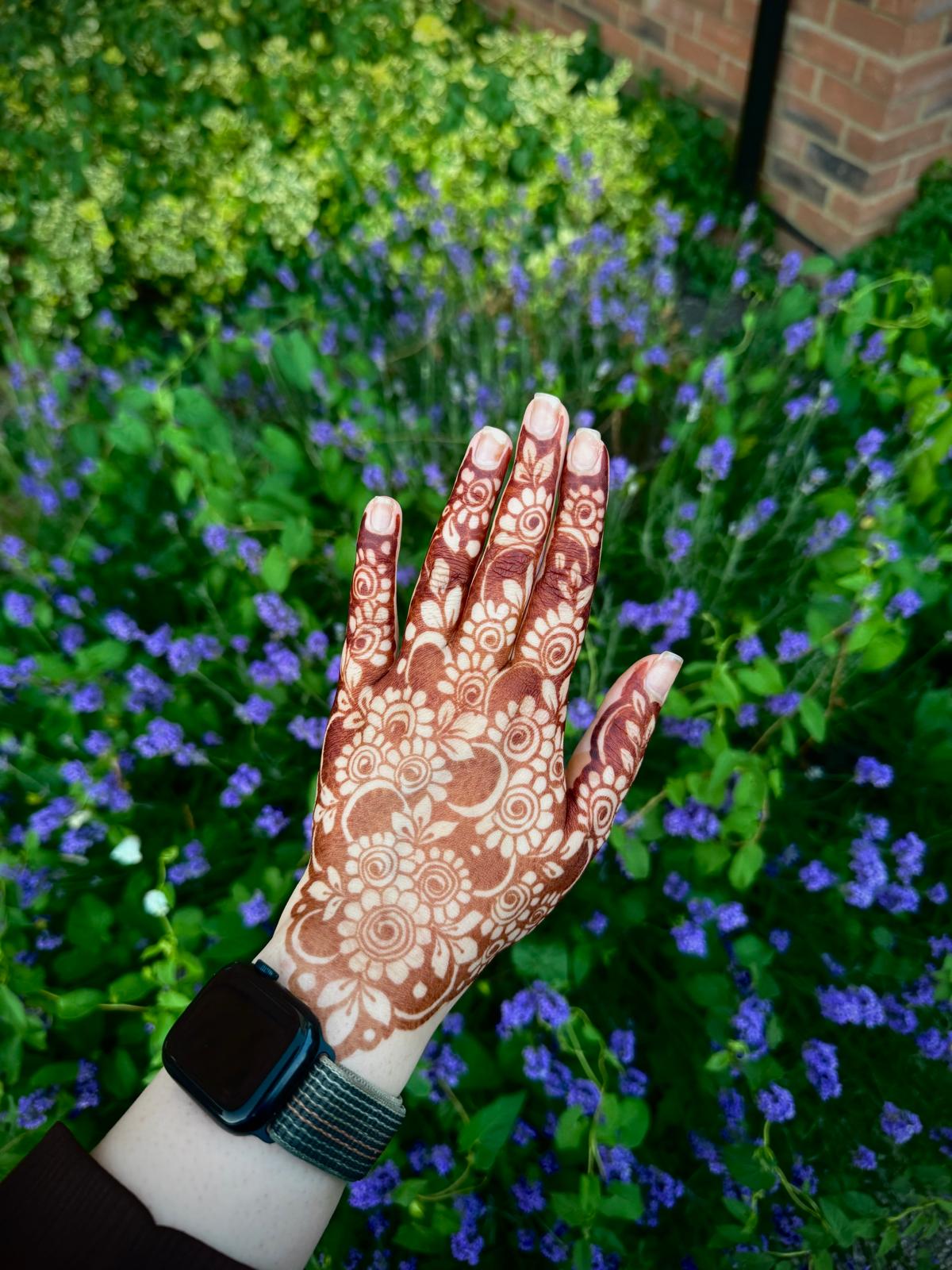 A hand with intricate brown henna patterns is raised in front of a blurred background of green foliage and purple flowers. A black smartwatch is visible on the wrist.