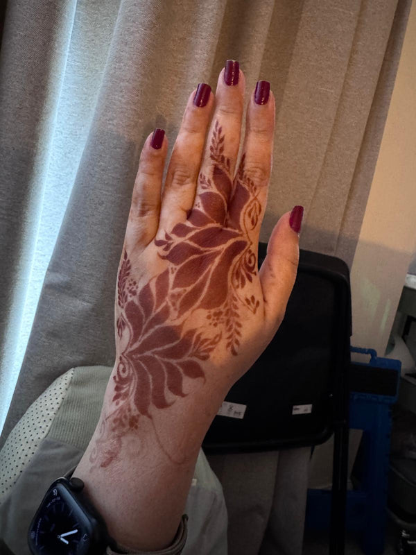 A close-up of a hand with a detailed brown henna floral design on the back of the hand, featuring dark red painted fingernails, set against a background of beige curtains and a black bag.